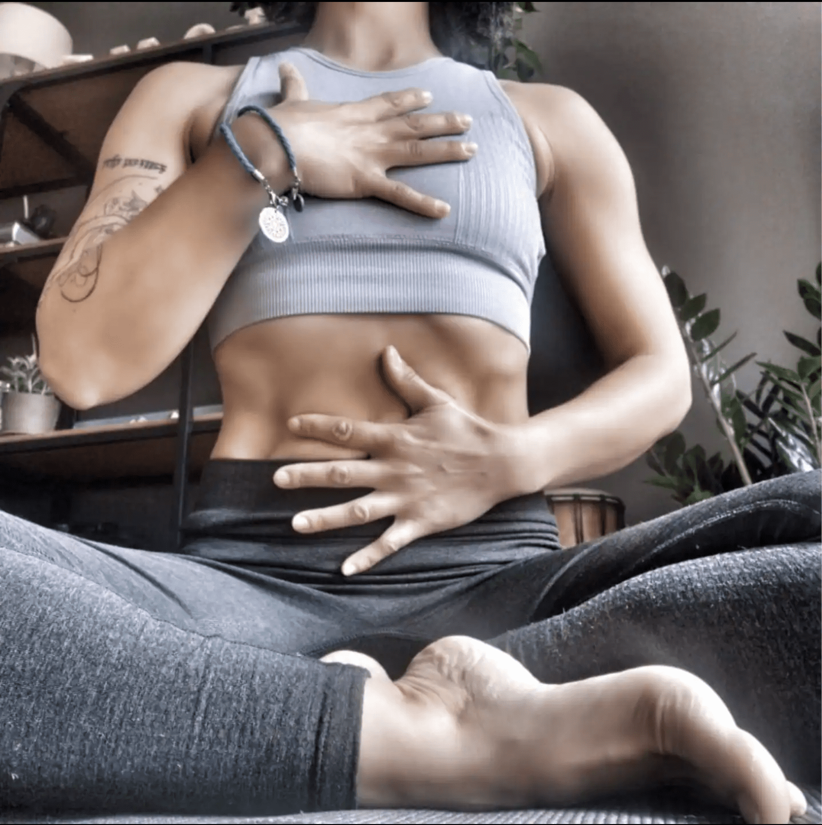 Wearing athletic wear, a person practices breathwork and mindfulness on a mat indoors during an Intuitional 1-1 Session, with plants and shelves visible in the background.